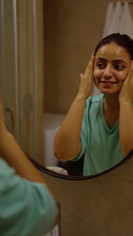 Happy-brunette-girl-with-Brown-skin-in-turquoise-pajamas-takes-care-of-her-body-skin-while-getting-ready-for-bed-in-the-evening-in-a-modern-apartment-in-front-of-a-mirror